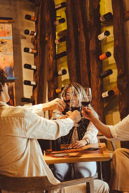 A group of people toasting with glasses of red wine at a table next to a rustic wooden wine rack.