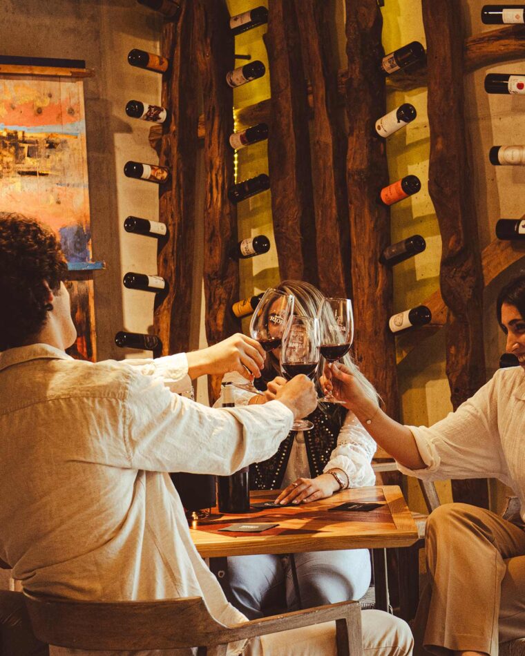 A group of people toasting with glasses of red wine at a table next to a rustic wooden wine rack.