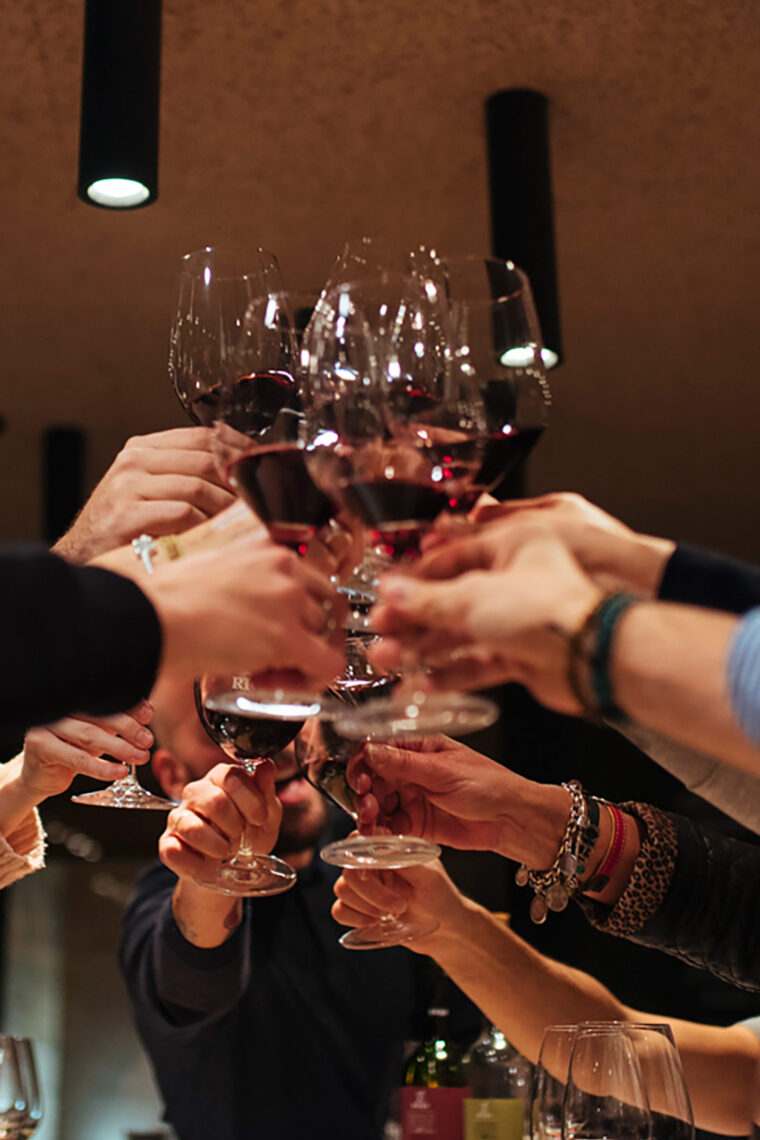 A large group of people clinking glasses of red wine together in a celebratory toast under modern lighting.