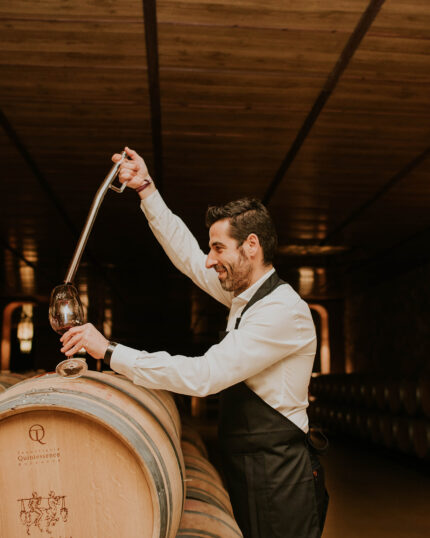 A winemaker in a cellar drawing red wine from a wooden oak barrel into a glass using a wine thief.