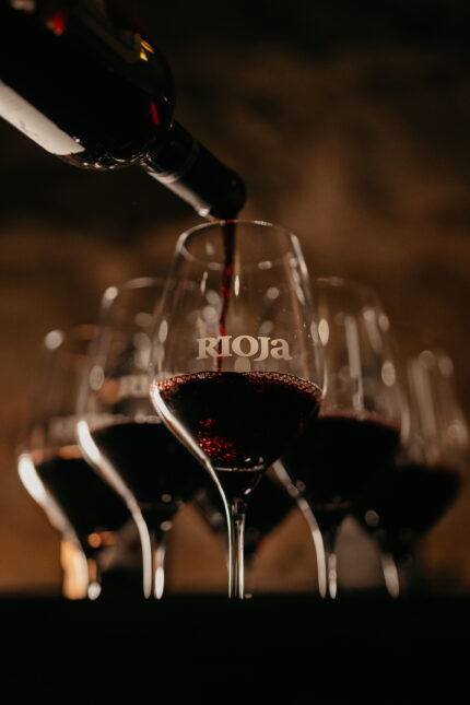 Red wine being poured from a bottle into a row of wine glasses, with the central glass.